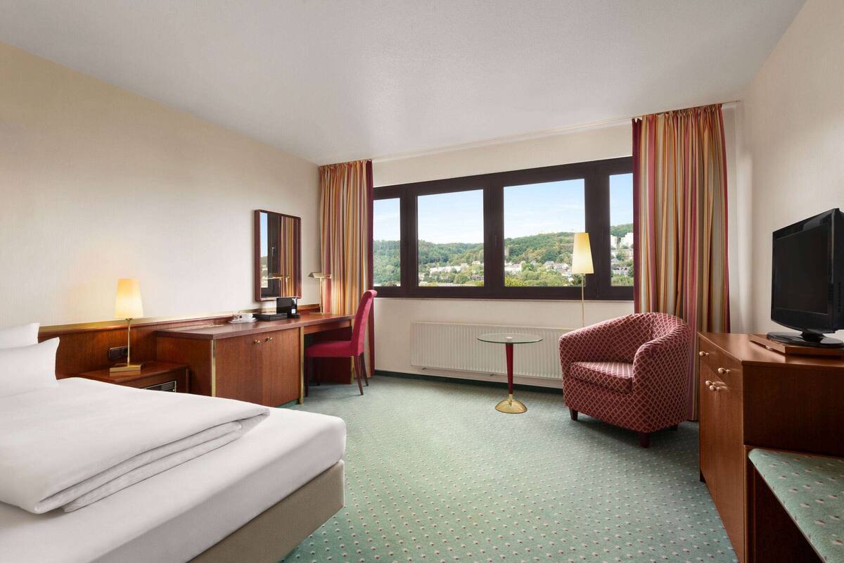 Habitación de hotel con cama, escritorio, silla, televisión y vistas al exterior a través de un amplio ventanal.