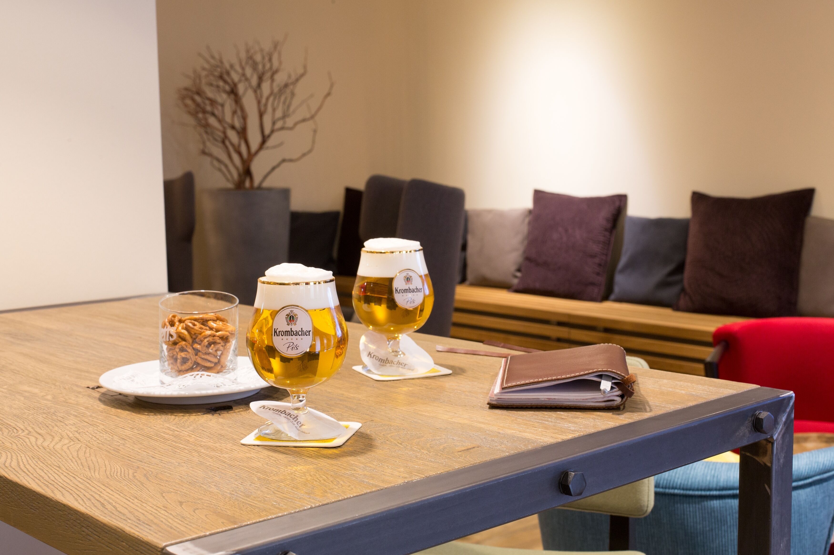 A table with two glasses of beer, a plate of pretzels, and a leather wallet on a wooden table in a modern room with a sofa and cushions in the background.