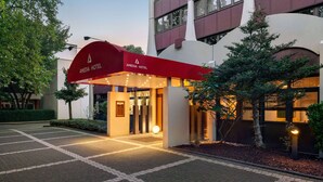 The exterior of the Amadia Hotel with a red awning, a paved entrance, and a landscaped area with trees and shrubs. - Amedia Siegen, Trademark Collection by Wyndham (Siegen)