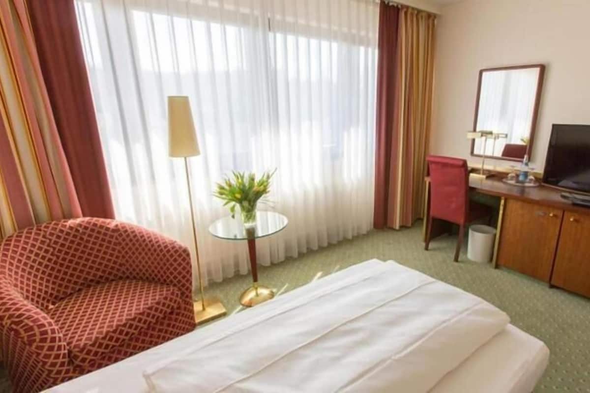 Una habitación de hotel con una cama, un escritorio con televisor, una silla, una mesita con una planta y cortinas.