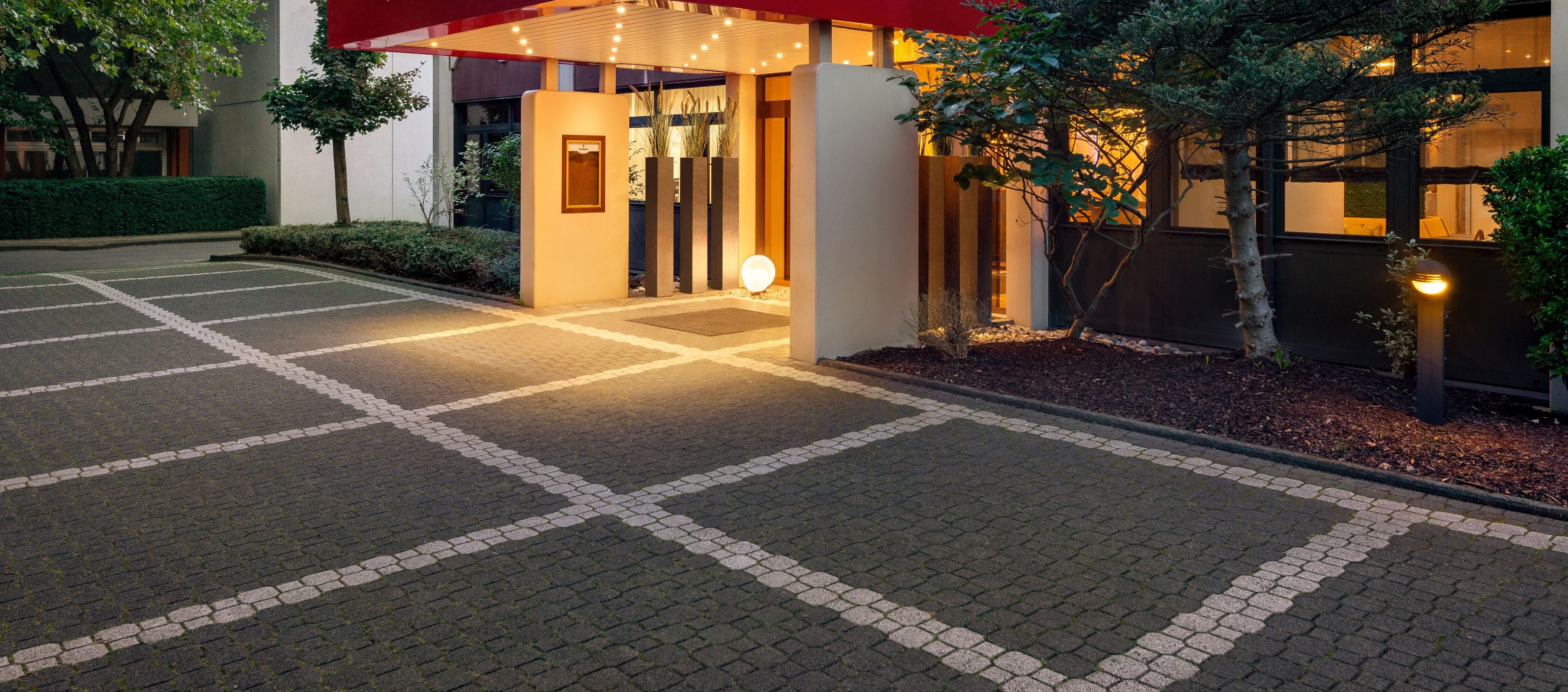 Un acceso pavimentado con un diseño geométrico, un toldo rojo y un camino bien iluminado que lleva a la entrada de un edificio.