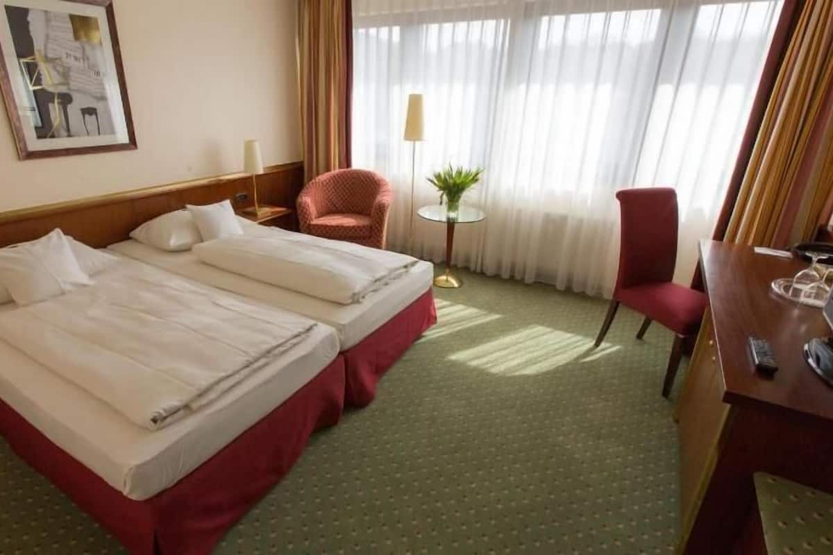 Habitación de hotel con dos camas, un escritorio con televisor, una silla, una mesita de noche con lámpara y una ventana con cortinas.