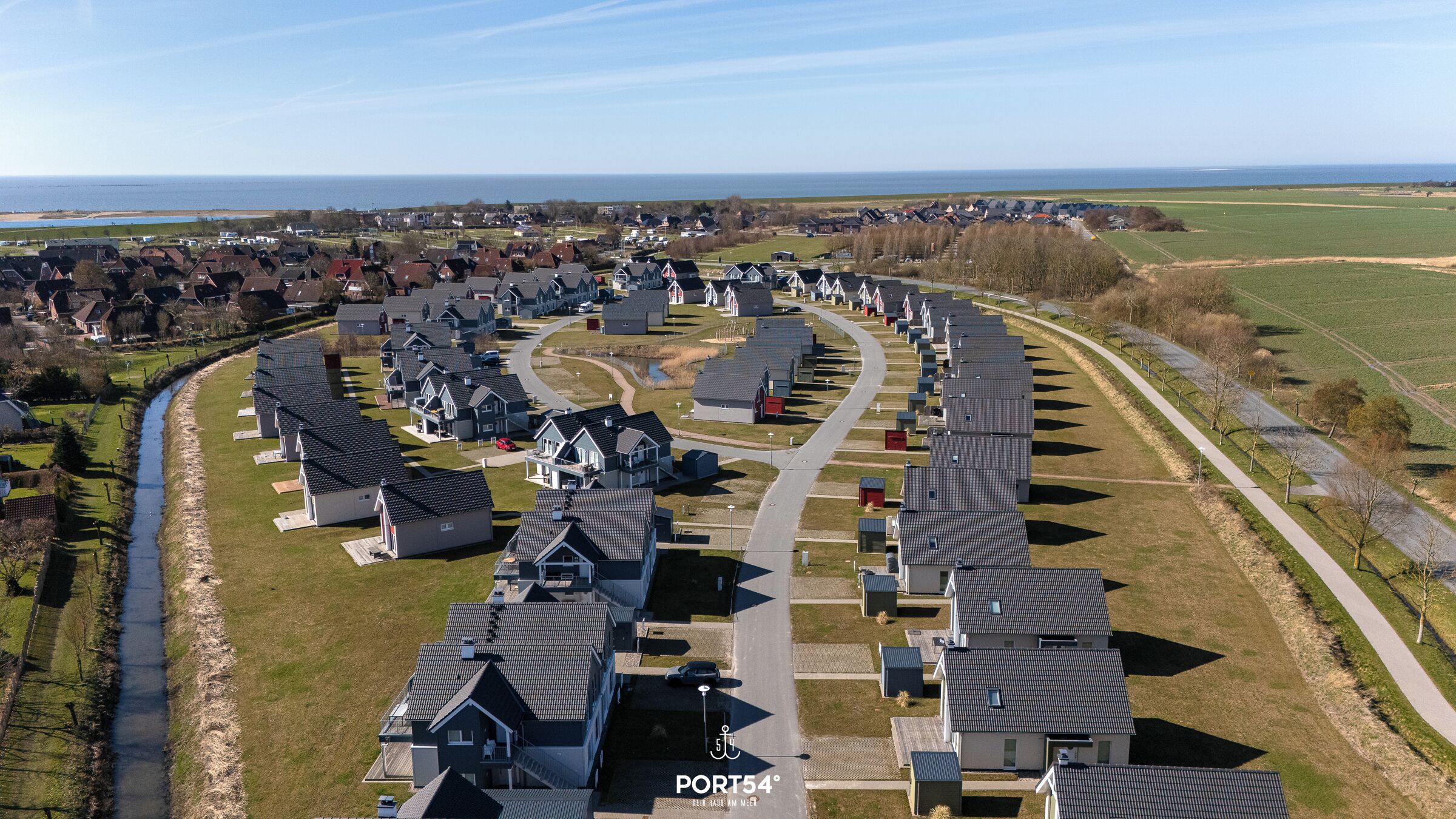 Ein Wohngebiet mit in einer Kurve angeordneten Häusern, einer Straße und einem Gewässer im Hintergrund.