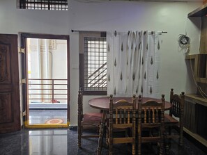 A room with a wooden dining table, chairs, a window with a view, a door with a metal gate, and a wall with a power outlet. - Private Two bedrooms flat in Hampi (Hampi)