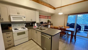 A modern kitchen with white cabinets, a stainless steel stove, and a large island with a sink. There is a dining area with a table and chairs, and a view of the outdoors through large windows. - Alpine Bliss Awaits: Beautiful Mountain Home with Hot Tub (Kimberley)
