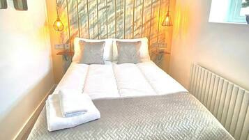 A neatly made bed with a textured white bedspread, two pillows, and a folded towel on the side. There are two wall-mounted lamps on either side of the bed, and a radiator is visible to the right.