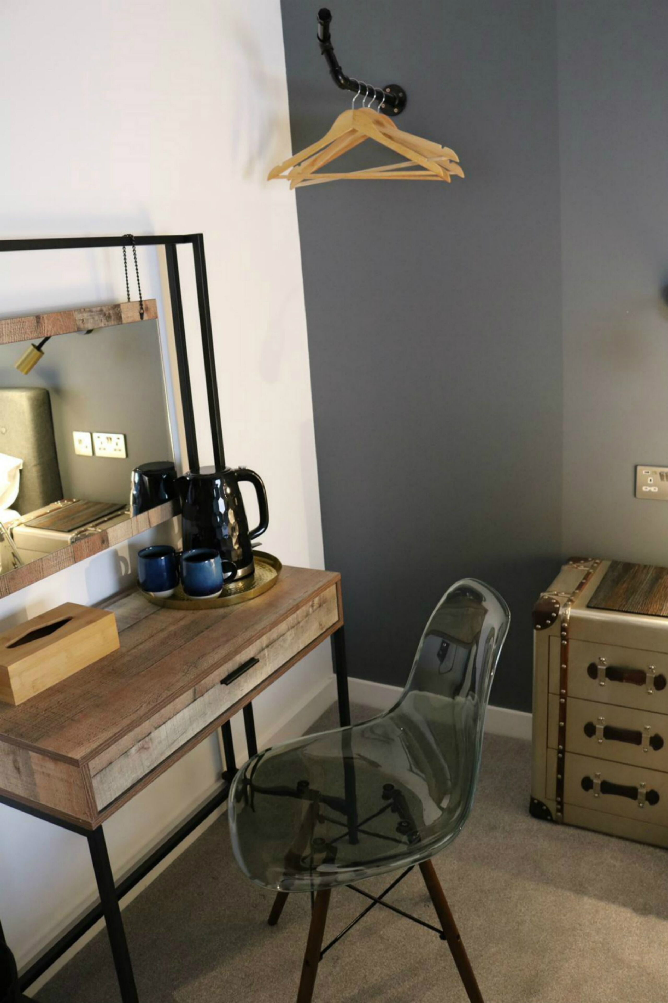 Una habitación pequeña con un escritorio de madera, una silla de vidrio, una percha para ropa y una maleta antigua.