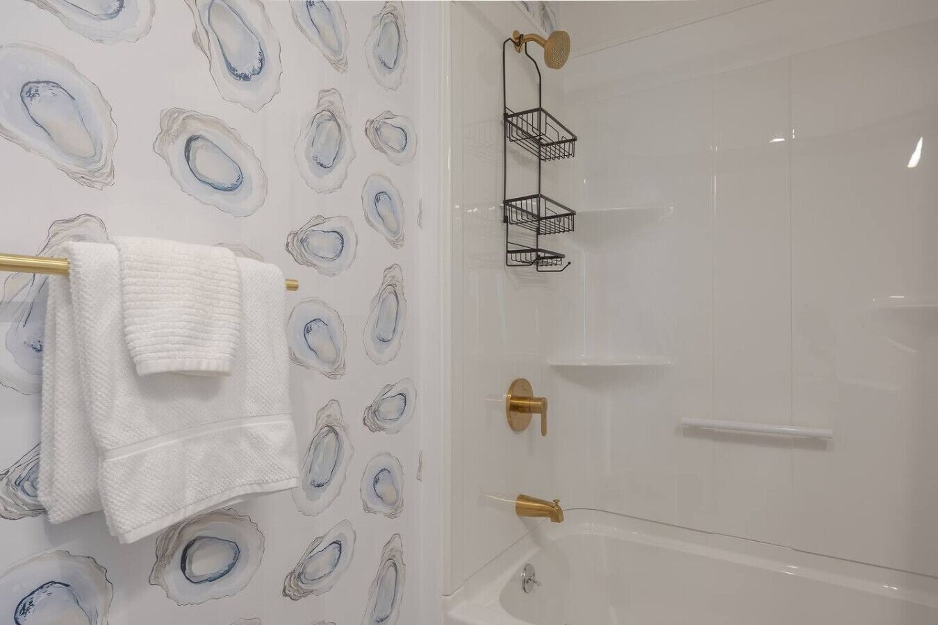 A bathroom with a white bathtub, a towel rack with a white towel, and a shower with a gold showerhead and black towel holders.
