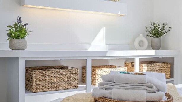 A modern bedroom with a white bed, a round ottoman, a wall-mounted shelf with decorative items, and a painting above the shelf. - Condo in the heart of town. (Bar Harbor)