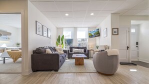 A spacious living room with a sofa, armchair, and coffee table, a dining area with a table and chairs, and a view of the outdoors through large windows. - Condo in the heart of town. (Bar Harbor)