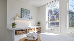 A bedroom with a bed, a bedside table with a lamp, a window with a view of houses, and a painting on the wall. - Condo in the heart of town. (Bar Harbor)