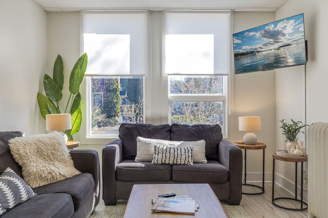 A living room with a grey sofa, a coffee table, two side tables with lamps, a large window with a view, and a painting on the wall.