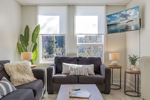 Ein Wohnzimmer mit grauem Sofa, einem Couchtisch, zwei Beistelltischen mit Lampen, einem großen Fenster mit Ausblick und einem Gemälde an der Wand.