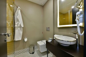 Un baño con mampara de ducha de vidrio, inodoro blanco, lavamanos con espejo y una toalla colgada en un perchero.