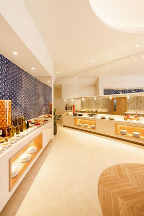 A modern hotel lobby with a central reception desk, a buffet area with various food items, and a seating area with a round wooden table. - Nova Maldives (Vakaru)