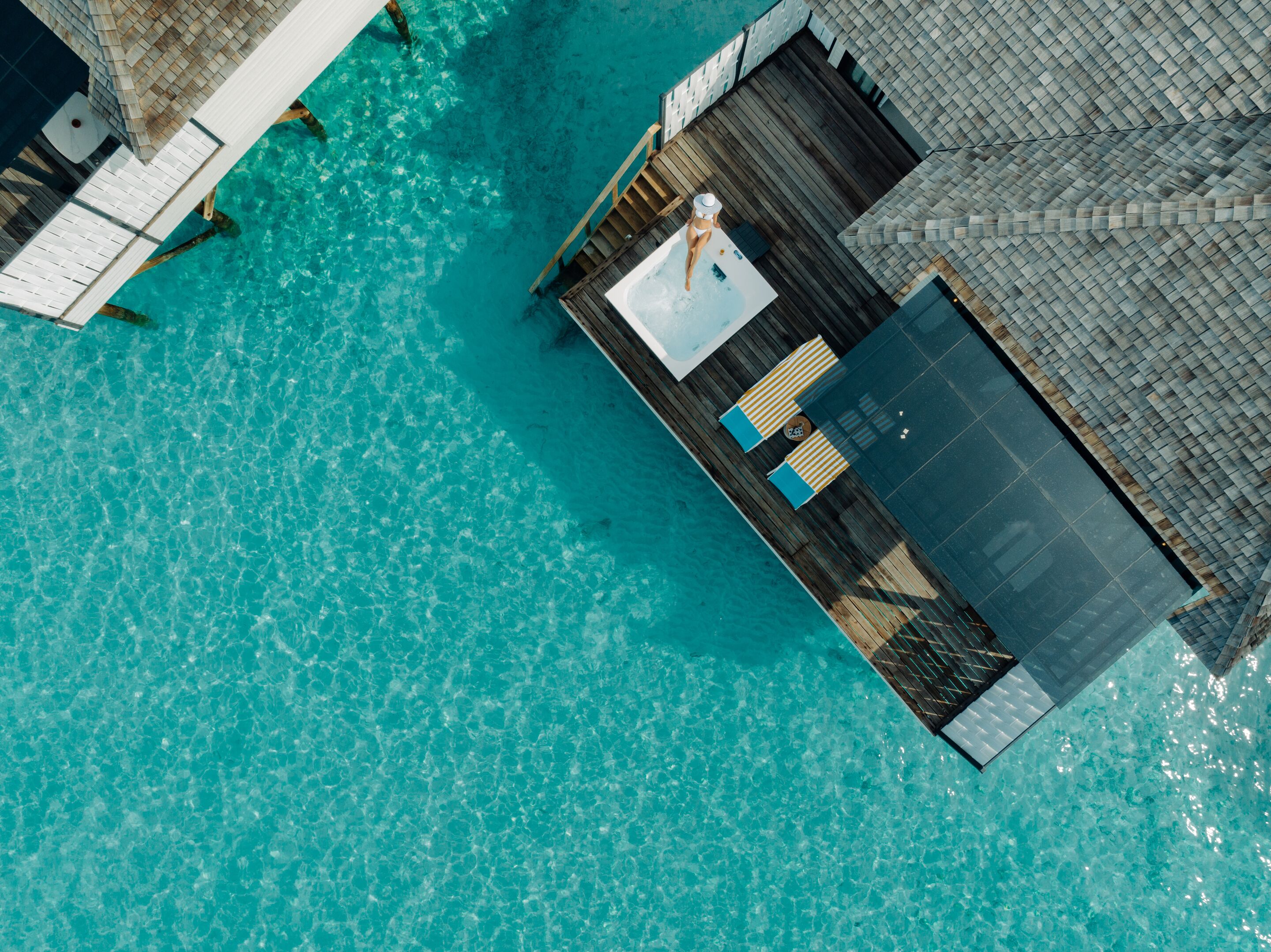 An aerial view of a resort with a clear turquoise pool, wooden deck, and a lounge area with striped cushions.