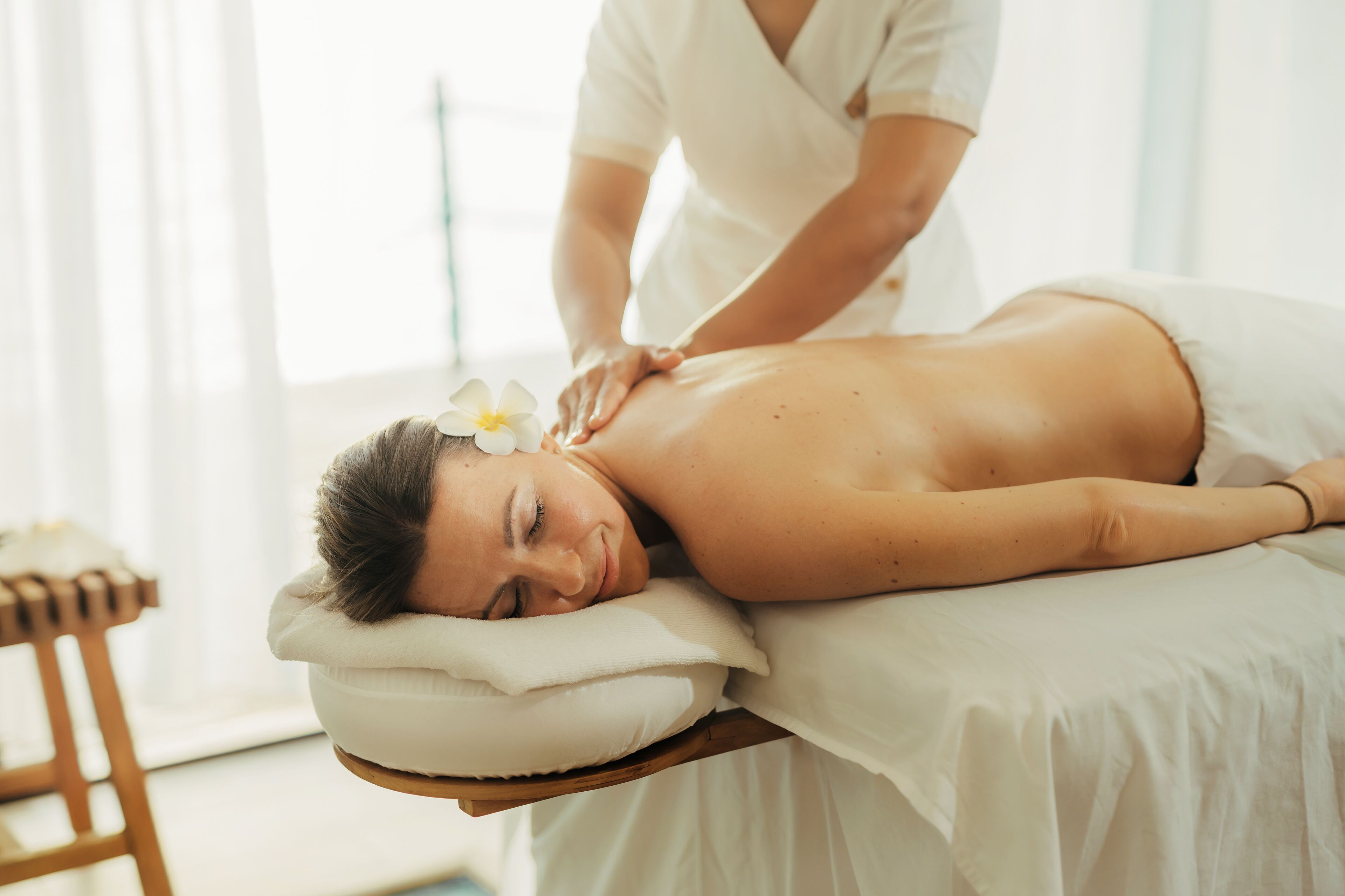 A person receiving a massage in a room with a wooden chair and a white bed.