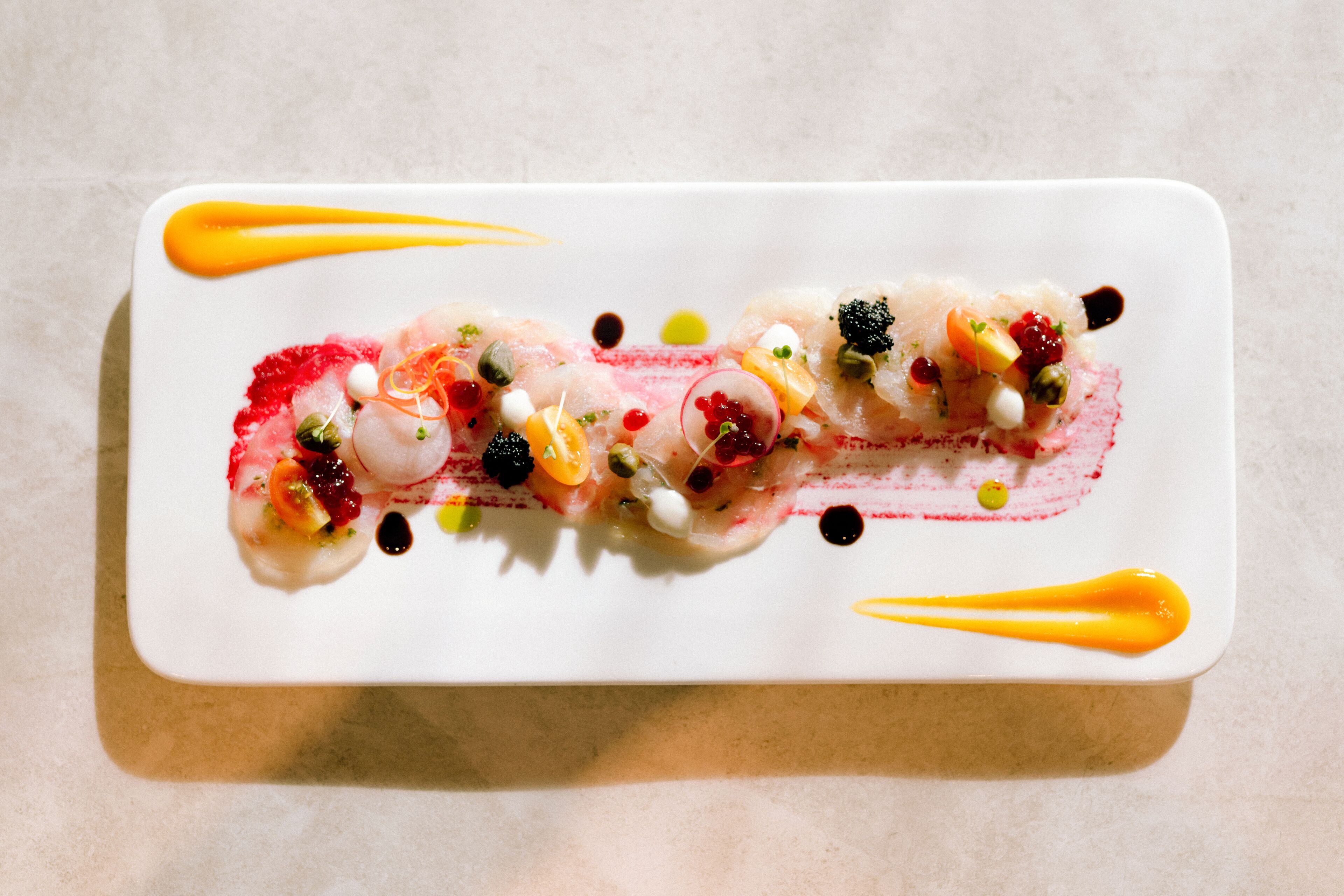 Une assiette rectangulaire blanche sur laquelle est présentée une préparation à base de fruits de mer, agrémentée de divers ingrédients colorés et de deux filets de sauce orange.