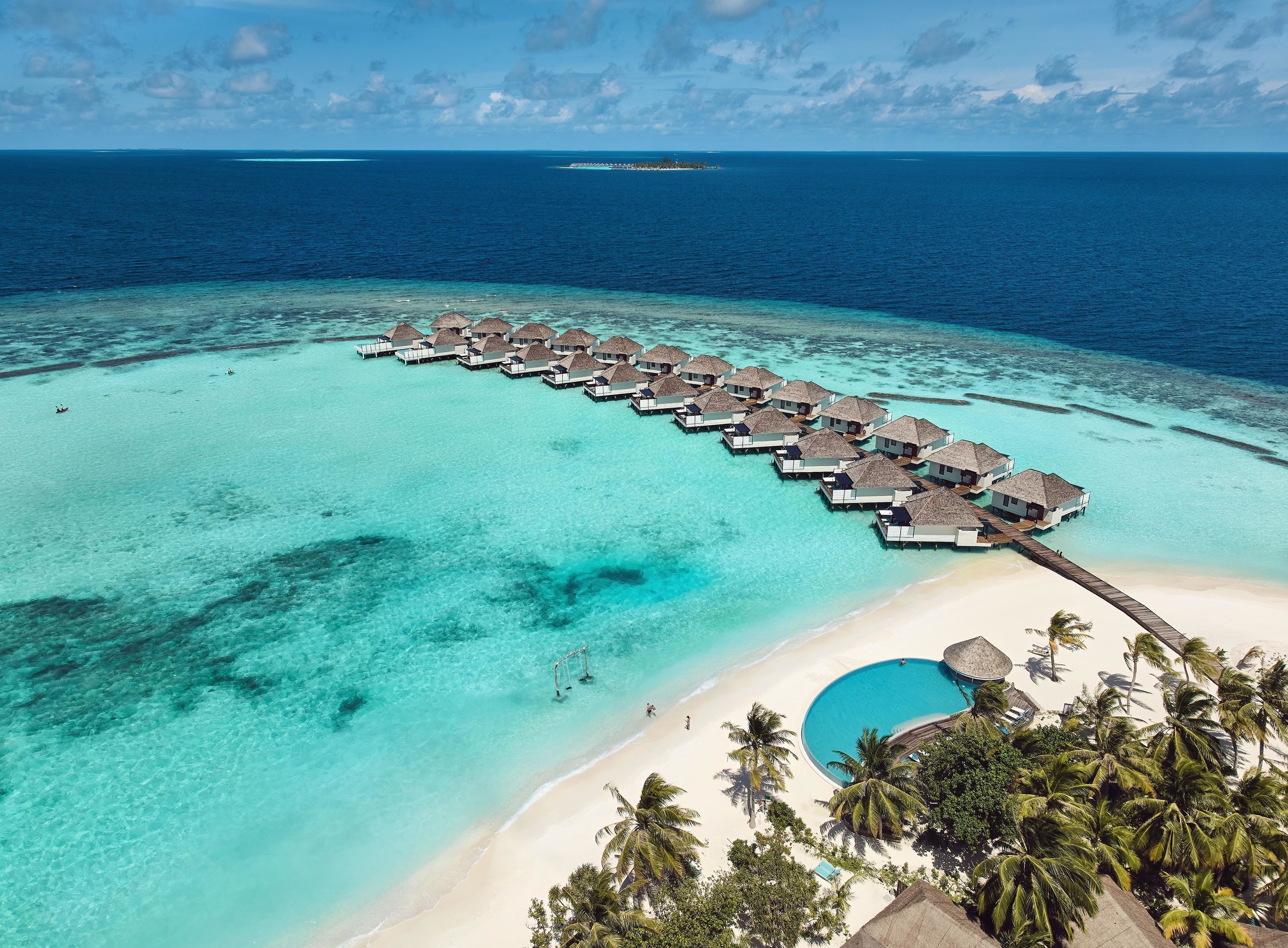 An aerial view of overwater bungalows connected by a wooden walkway, a clear turquoise lagoon, and a sandy beach with palm trees.