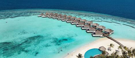 An aerial view of overwater bungalows connected by a wooden walkway, a clear turquoise lagoon, and a sandy beach with palm trees.