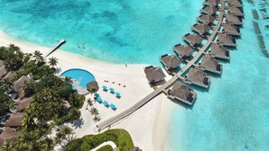 Vista aerea di un resort con bungalow sull'acqua, una spiaggia sabbiosa e un mare turchese cristallino.