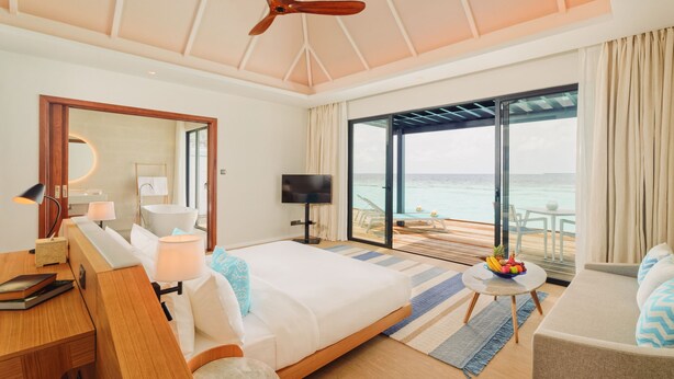 A modern hotel room with a large bed, a desk, a TV, and a balcony with a view of the ocean. - Nova Maldives (Vakaru)