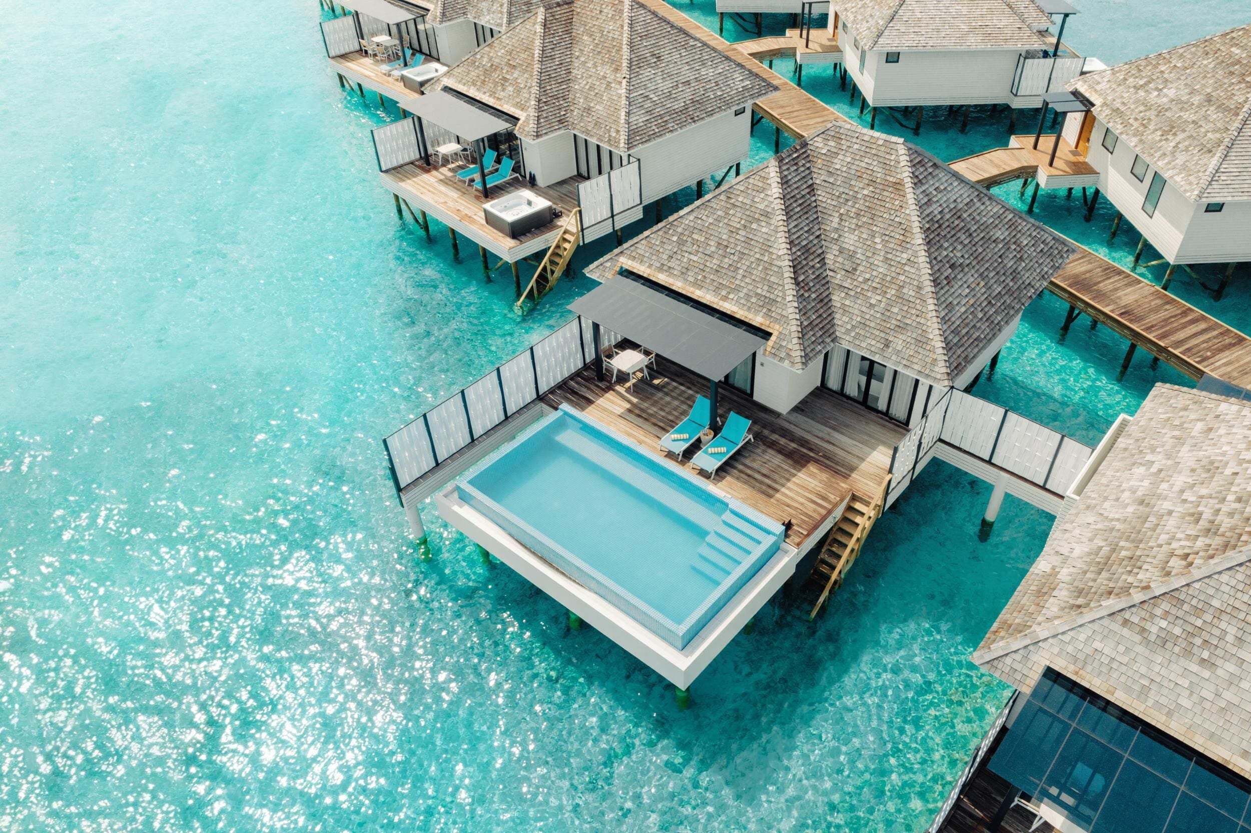 An aerial view of overwater bungalows with a swimming pool, wooden deck, and lounge chairs.