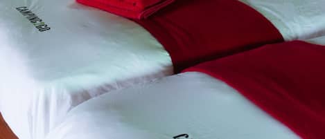 Una cama bien hecha con ropa de cama blanca y detalles rojos, incluyendo una toalla roja y almohadas. La cama tiene un cabecero de madera y está ubicada frente a una ventana con vistas a la vegetación.