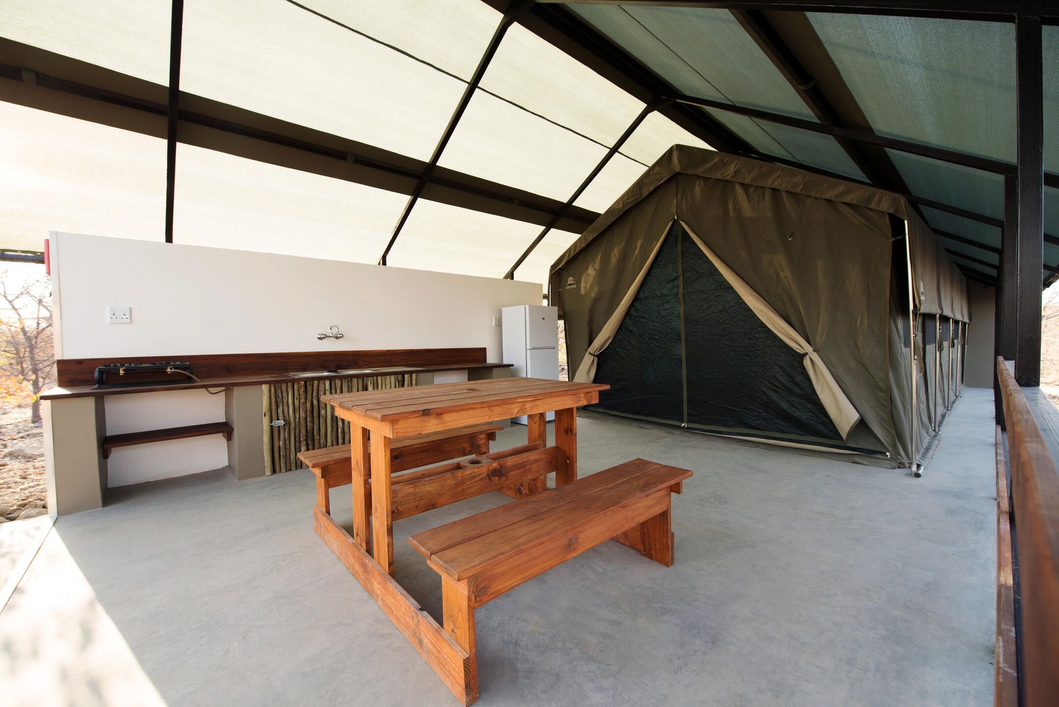 A covered outdoor kitchen area with a wooden table and bench, a tent, and a large window.