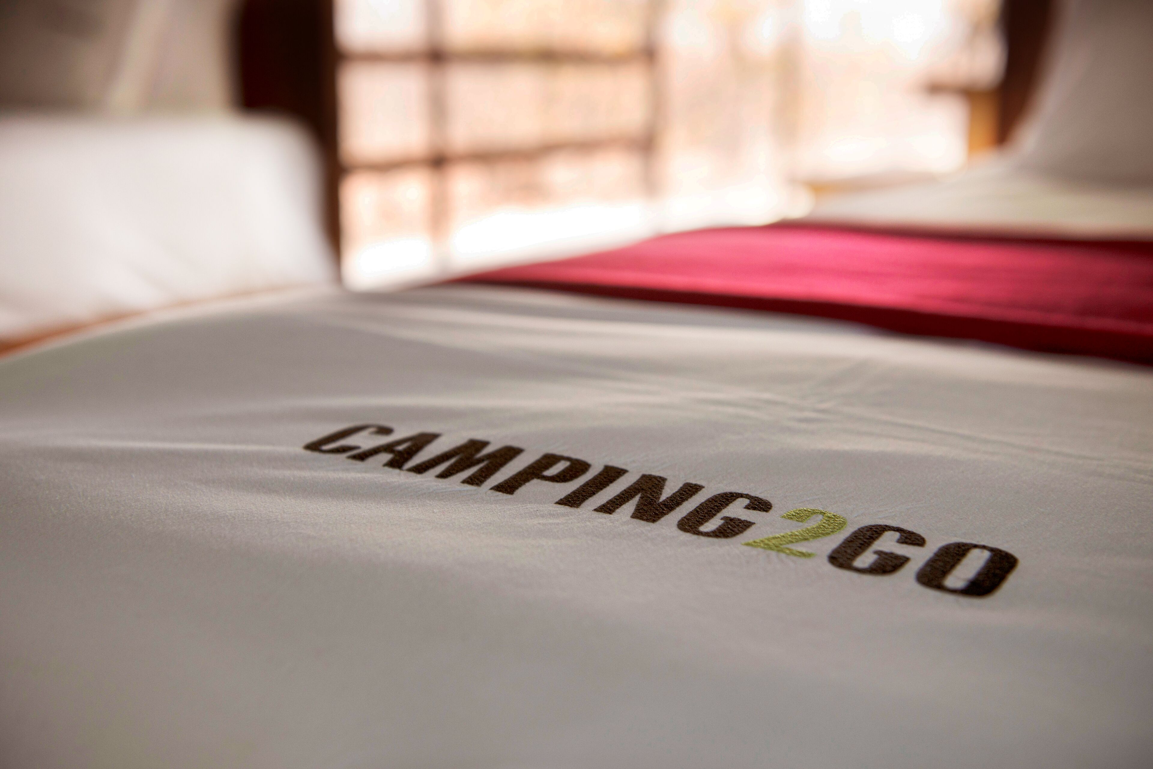 A bed with a white sheet and a red blanket. The text "CAMPING 2GO" is visible on the sheet.