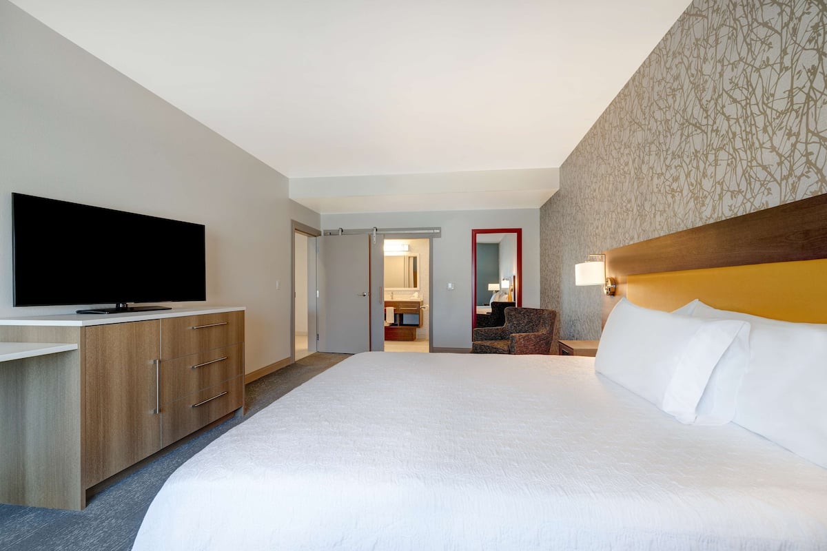 Habitación de hotel con una cama grande, un televisor sobre un tocador de madera, una silla y un baño visible a través de una puerta abierta.