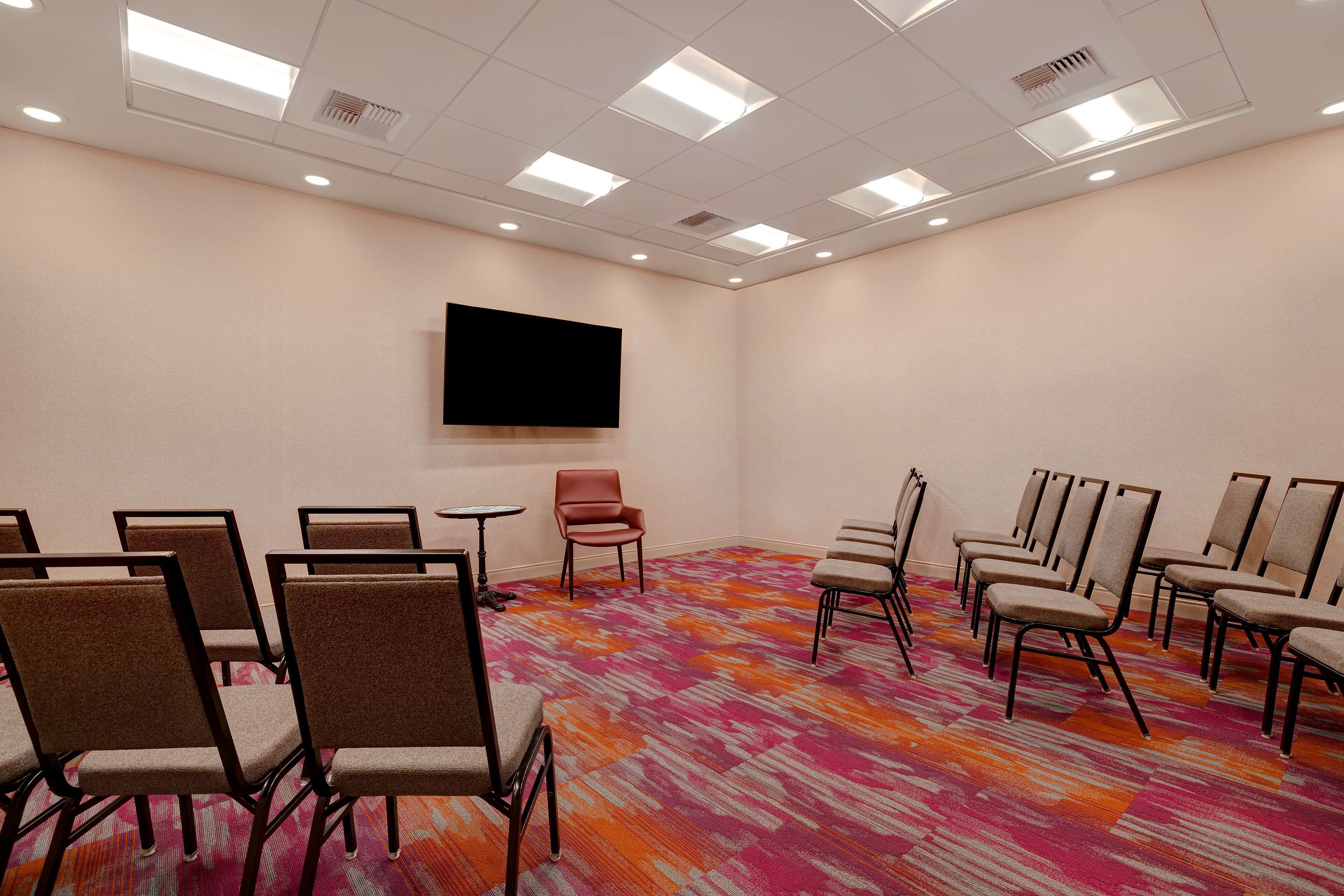 Un salón de conferencias con filas de sillas, un televisor de pantalla plana en la pared y una alfombra colorida.