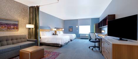 A hotel room with a sofa, two beds, a desk, a television, and a patterned rug.