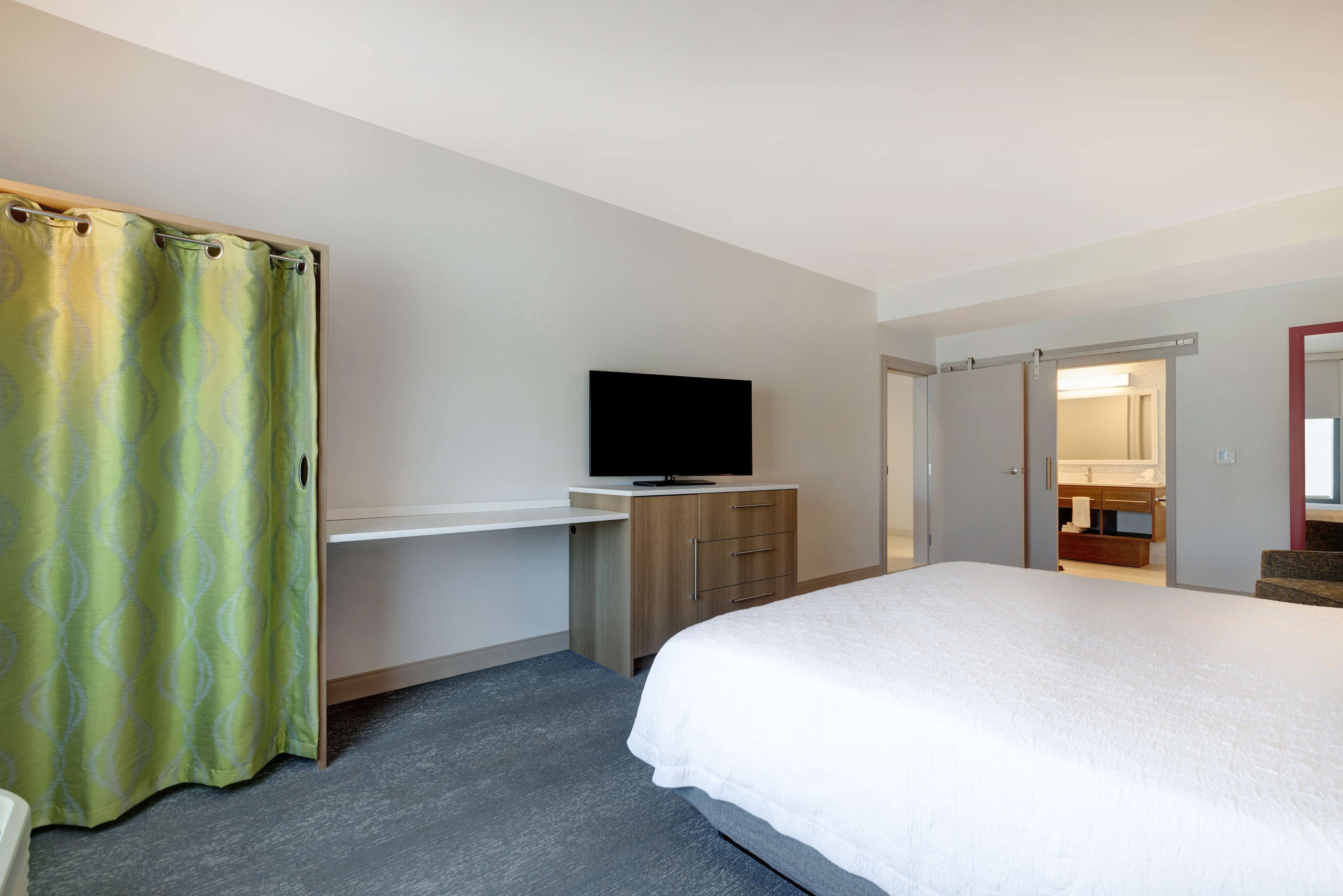 A hotel room with a bed, a desk with a flat-screen TV, a bathroom with a mirror and sink, and a green curtain.