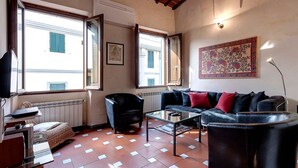 Ein Wohnzimmer mit einer Couch, Sesseln, einem Couchtisch und einem Fernseher. Es gibt Fenster mit Rollläden, eine Heizung und ein gerahmtes Bild an der Wand.