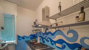 A kitchen with a gas stove, a sink, and a window with blinds. There are shelves with jars and bottles above the stove. - Sciacchetrà Apartment with AC (Monterosso al Mare)