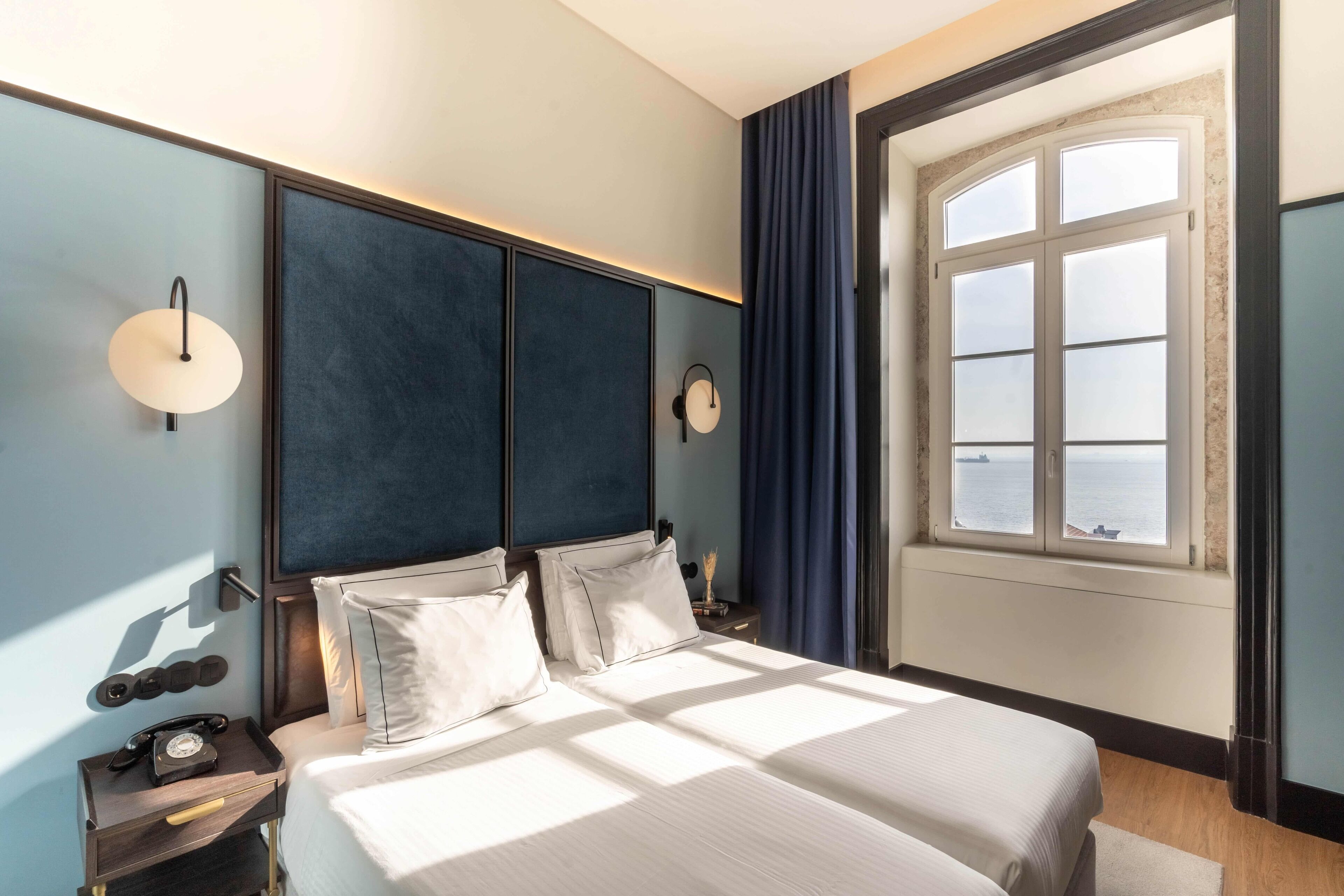 A hotel room with a large bed, bedside tables, a nightstand with a telephone, and a window with a view of the sea.