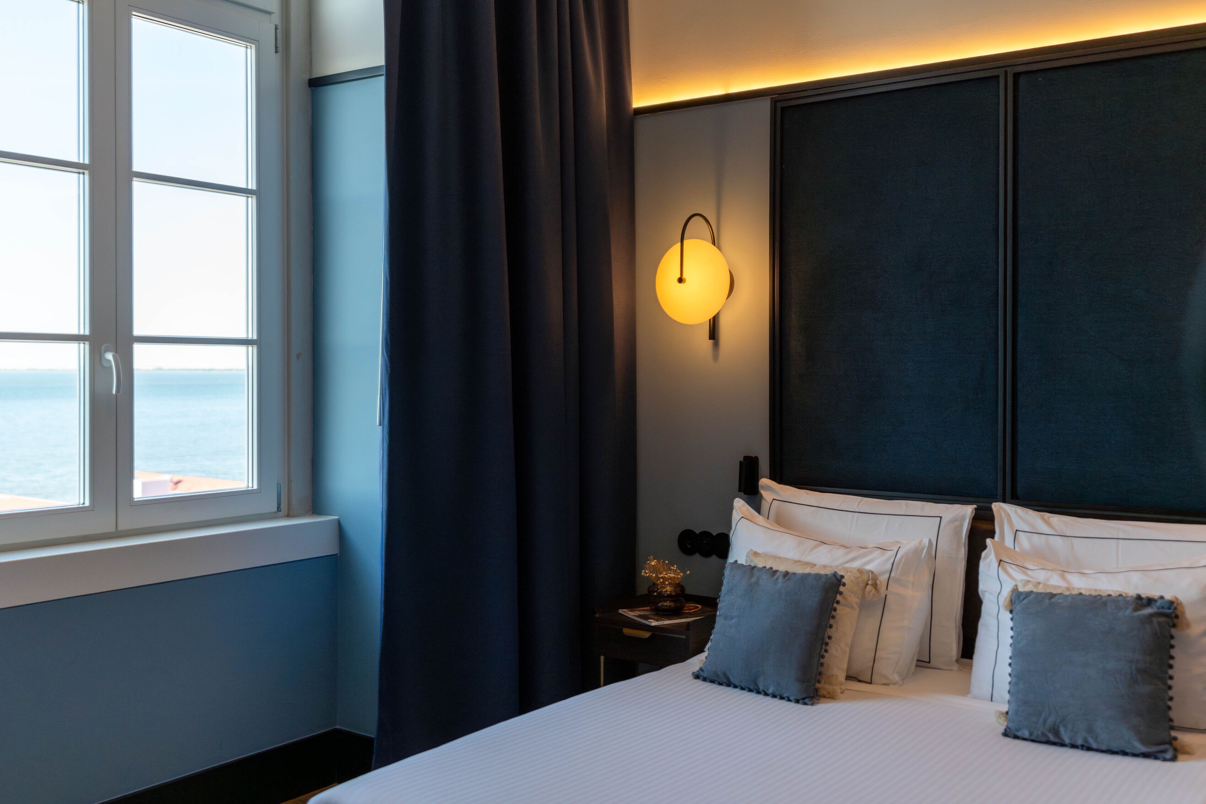 Habitación de hotel con una cama grande, una mesita de noche, una lámpara de pared y vistas al mar a través de una ventana.