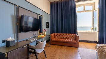 A modern hotel room with a large flat-screen TV, a leather sofa, a desk with a chair, and a view of the sea through a large window.