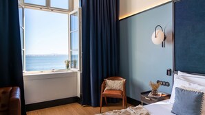 A hotel room with a large window, a bed with white bedding, a wooden chair, and a nightstand with a lamp. - The Editory Riverside Hotel, an Historic Hotel (Lisbon)