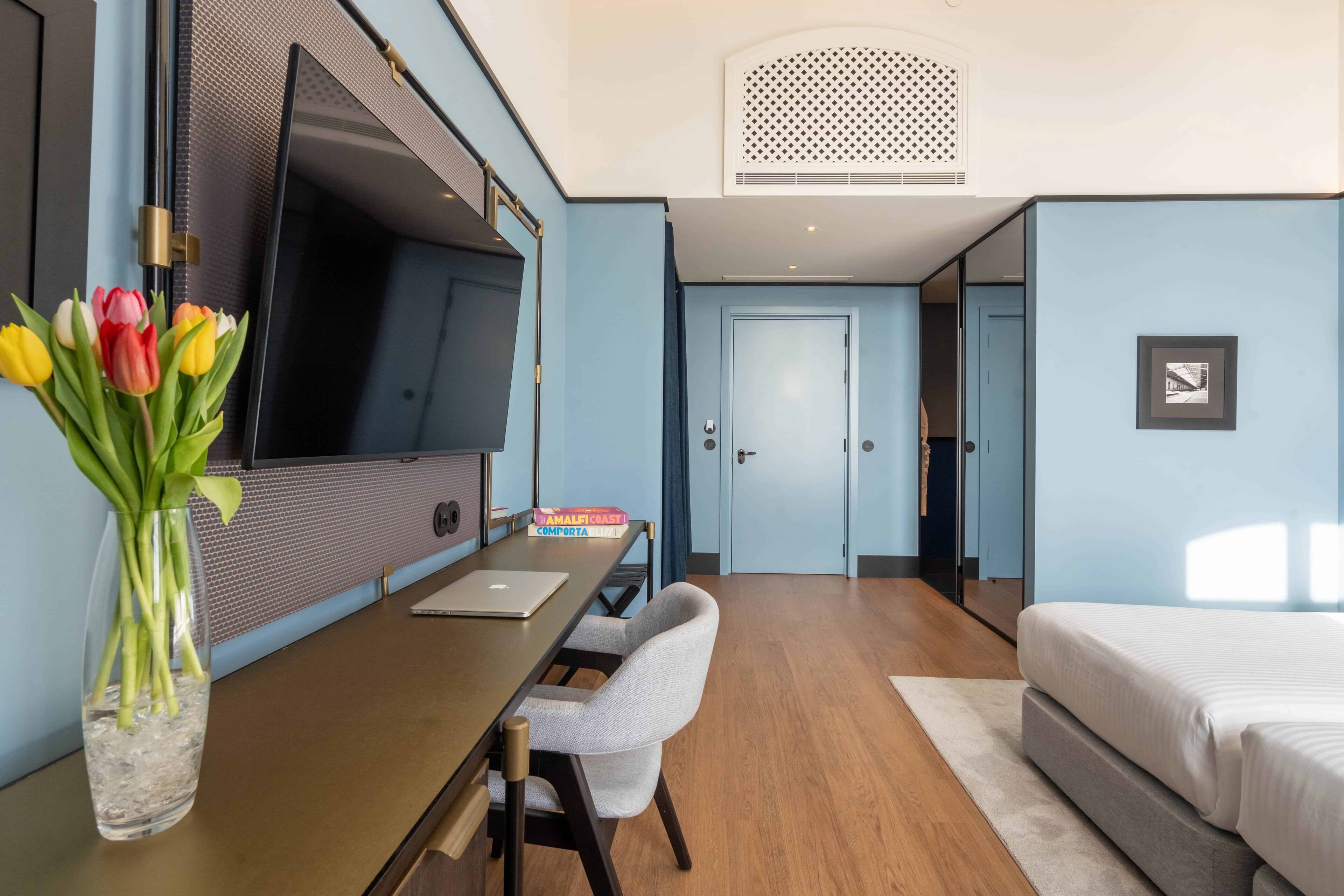 A hotel room with a bed, a desk with a laptop, a TV, and a vase of tulips.