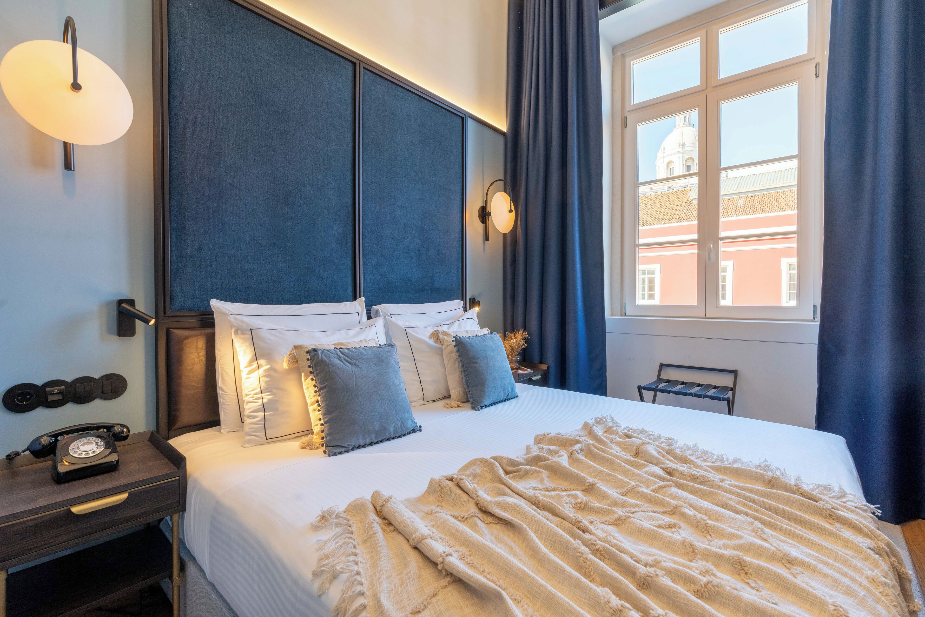 A hotel room with a large bed, a bedside telephone, a nightstand, and a window with curtains.