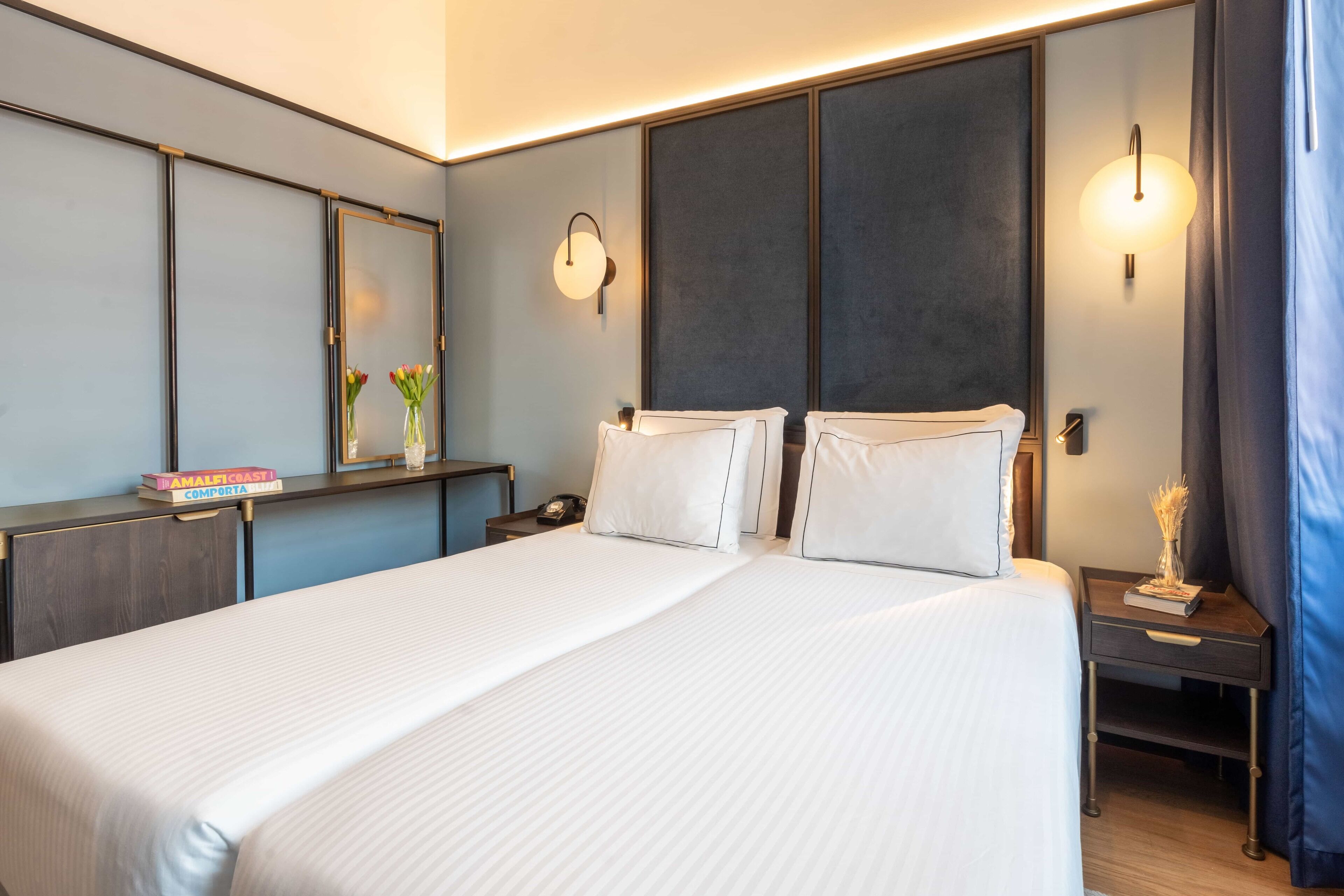A modern hotel room with a large bed, bedside tables, a mirror, and wall-mounted lights.