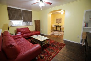 A living room with a red leather couch, a wooden coffee table, and a patterned rug. There is a ceiling fan, a window with blinds, and a door leading to another room.