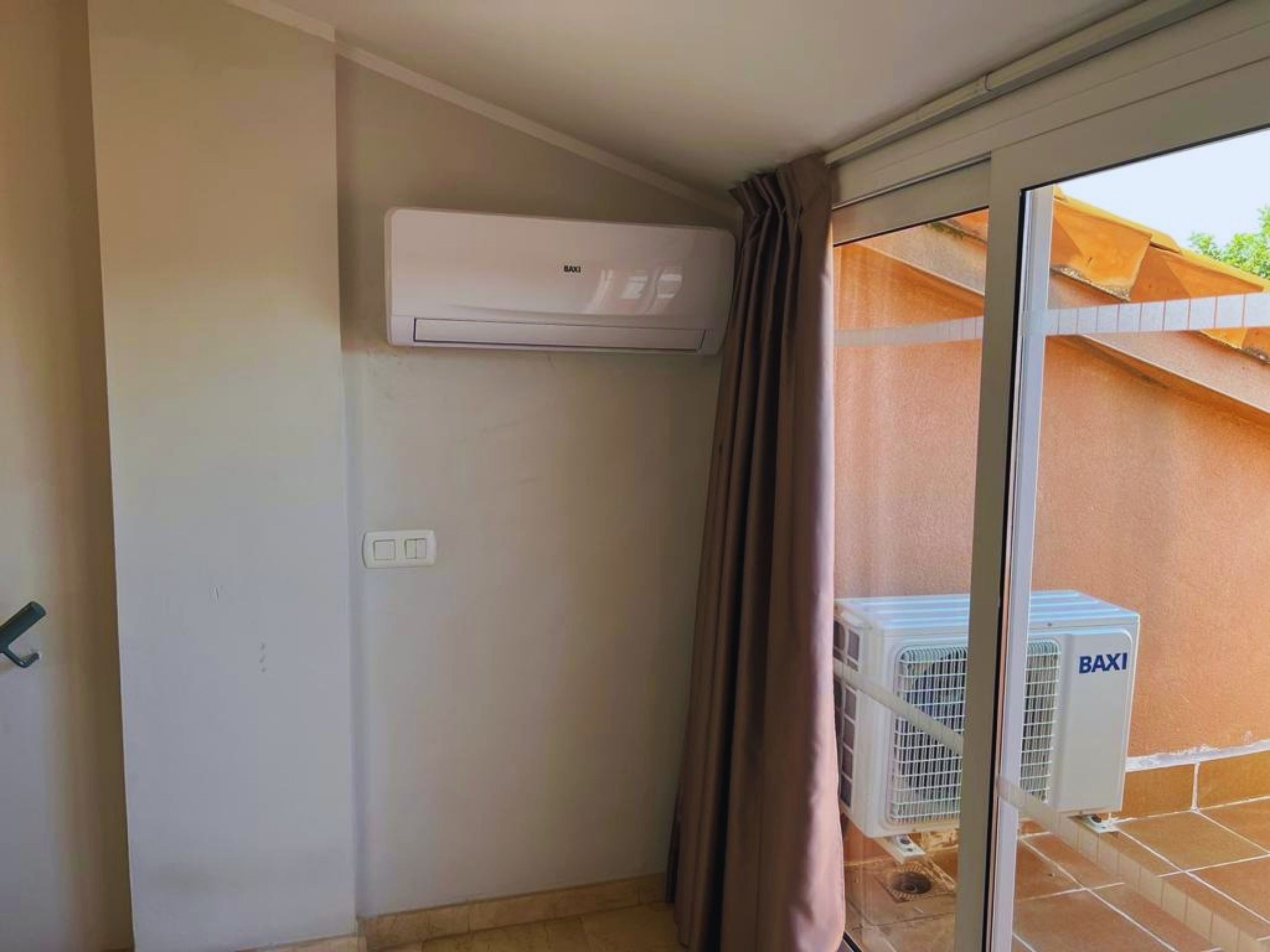 Una habitación con puerta de vidrio corredera, aire acondicionado de pared y ventana con vista a un edificio.