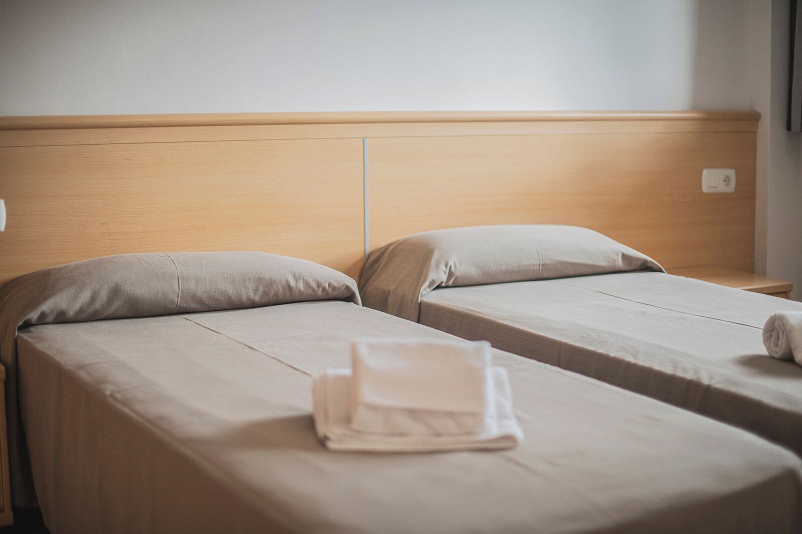 Two single beds with wooden headboards and beige bedding, a small table, and a folded towel on the floor.