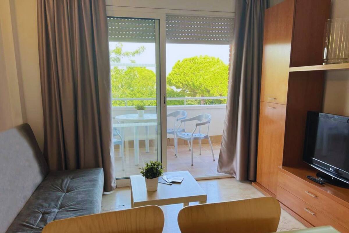 A modern living room with a sofa, a coffee table, and a TV. There is a balcony with a table and chairs, and a view of greenery.