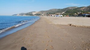 A wide sandy beach with footprints leading to the water, a clear sky, and distant hills. - Marsol Apartamentos Turisticos (Castelldefels)
