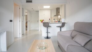 A modern living room with a sofa and two bar stools, a wooden coffee table, and a kitchen area in the background. - Marsol Apartamentos Turisticos (Castelldefels)