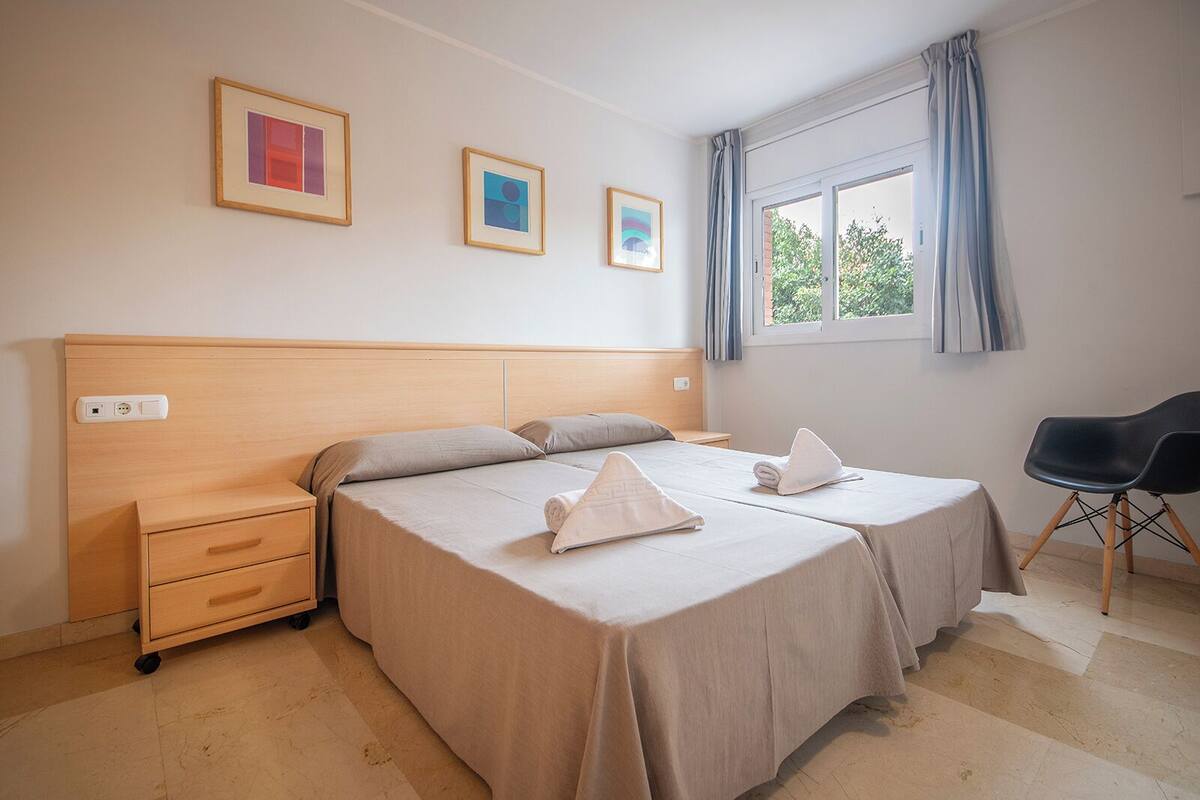 A hotel room with a wooden headboard, two beds, a desk, and a chair. There are three framed pictures on the wall and a window with curtains.